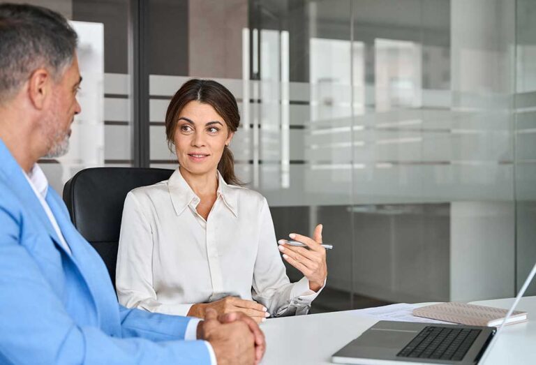 A police officer speaking with a person seated in a small interview room, both looking serious and focused.